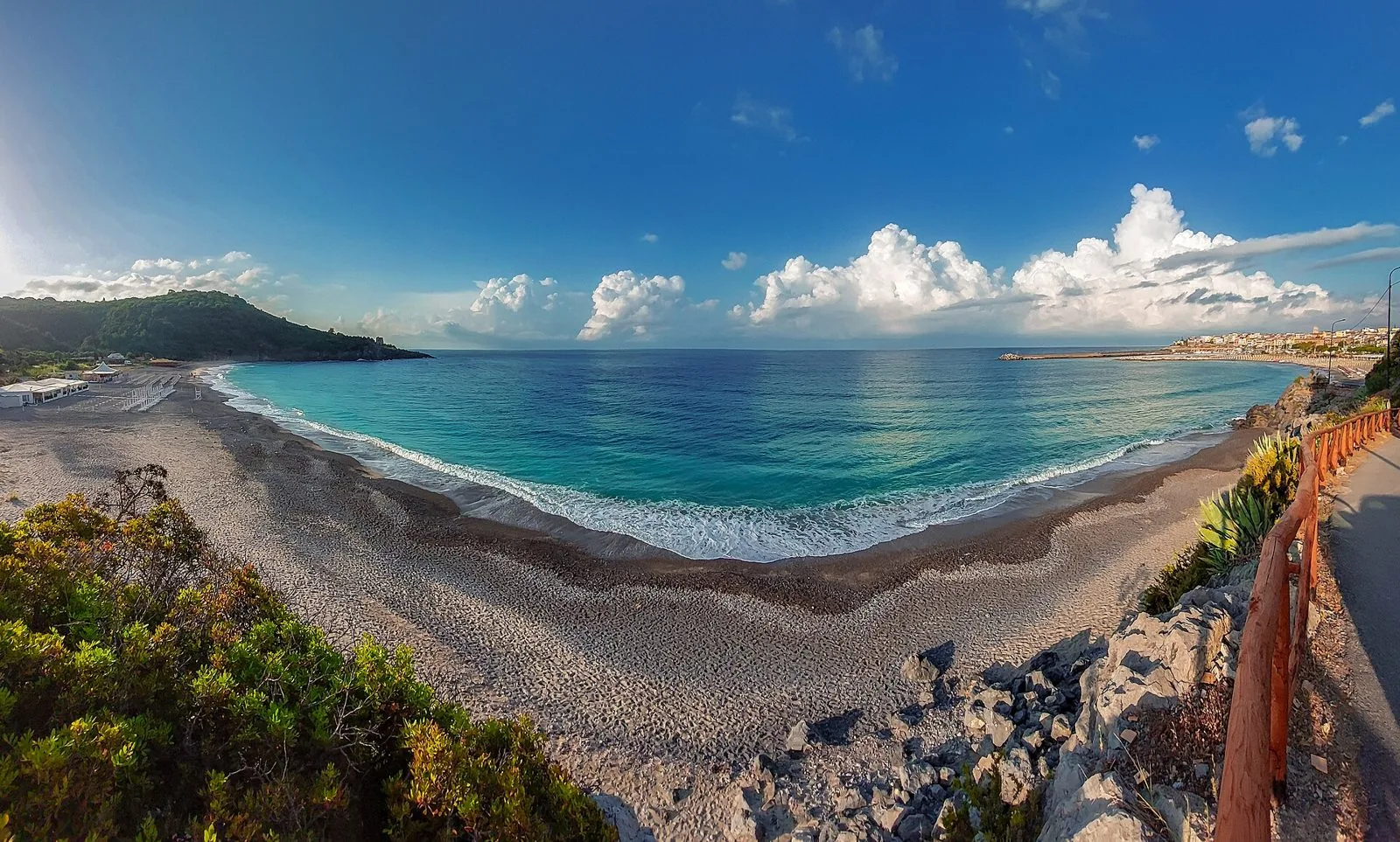Ampia spiaggia di ciottoli chiari a Marina di Camerota con mare turchese, promontorio verde a sinistra e il paese con il molo a destra